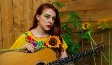 Holly Clausius promo photo. Holly is sitting amongst sunflowers wearing a bright yellow top and yellow makeup while holding her guitar