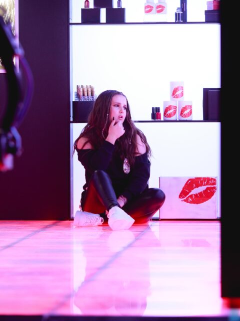 Victoria Anthony sitting on the floor on set of her music video in a scene where she is in a makeup department. The colours of the lights around her are pink and white. She is wearing all black and we can see a camera being pointed at her.