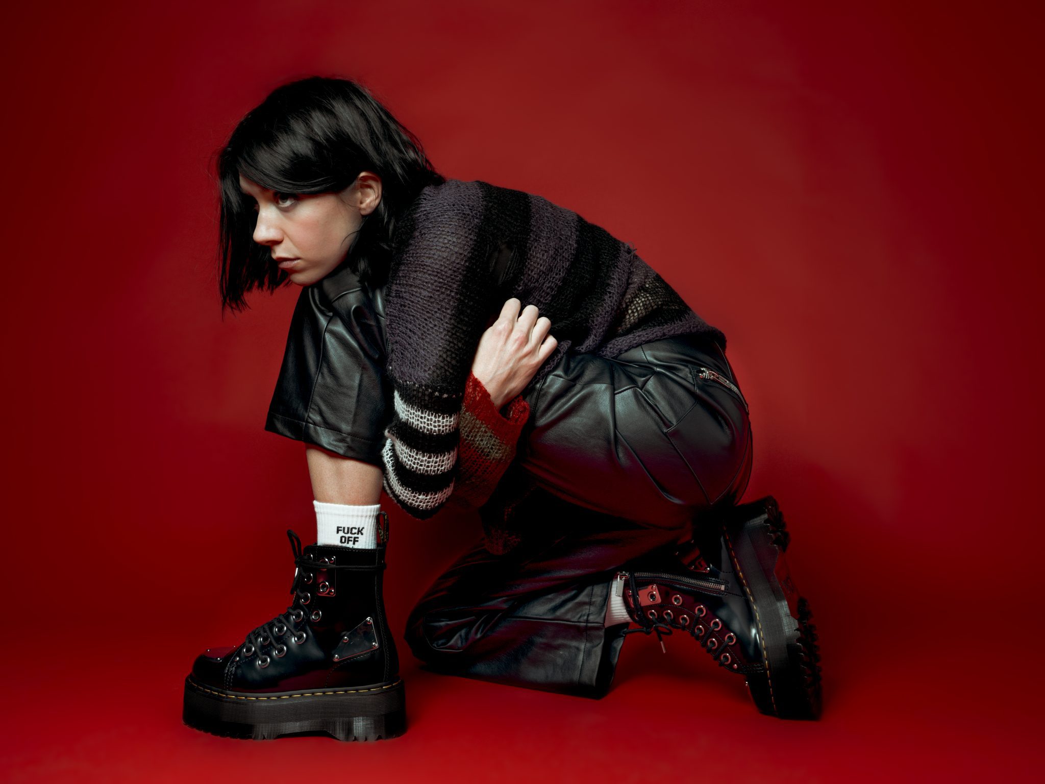 K Flay press photo. K Flay is sitting on the floor wearing all black in front of a red backdrop.