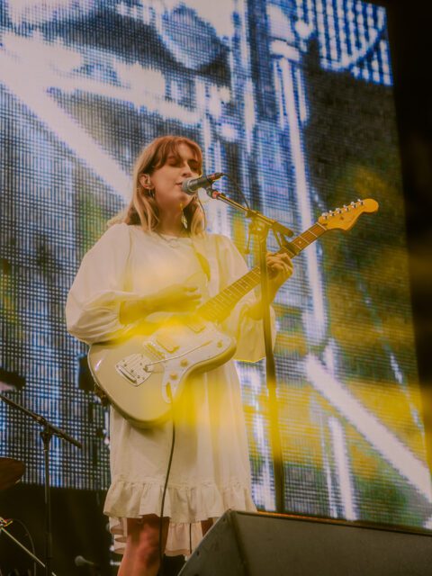 Singer songwriter Ellis is performing on stage. She is wearing a white dress and singing into a microphone. She is playing a green and white electric guitar.