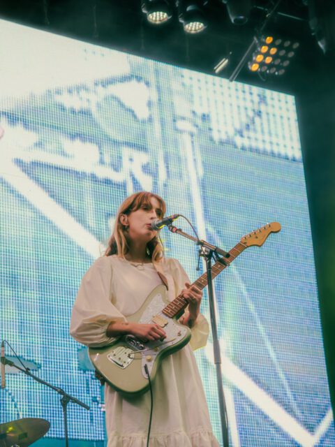Singer songwriter Ellis is performing on stage. She is wearing a white dress and singing into a microphone. She is playing a green and white electric guitar.