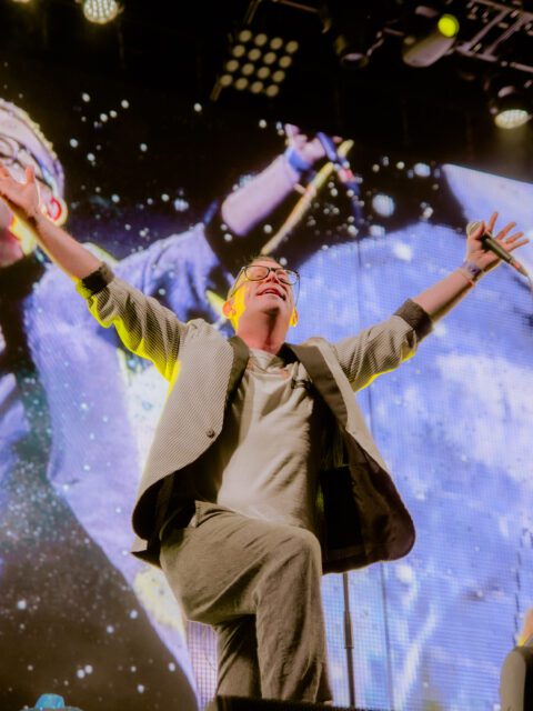 Torquil Campbell of the band Stars on stage with his arms thrown up in the air above him. He is holding a microphone in one hand and smiling.