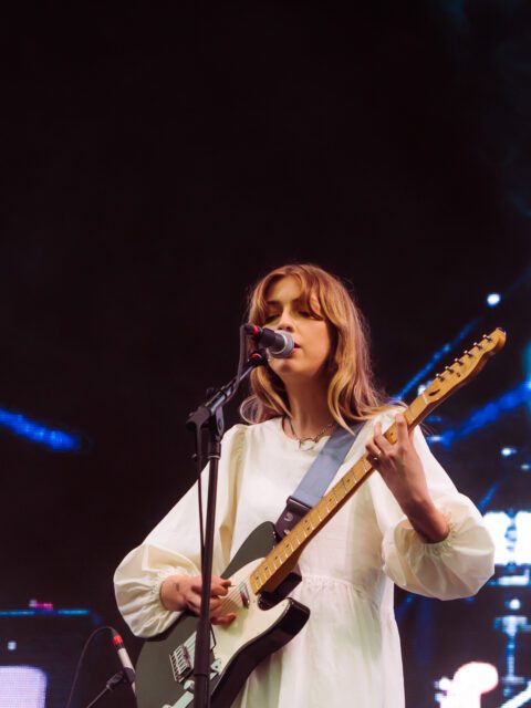 Singer songwriter Ellis is performing on stage. She is wearing a white dress and singing into a microphone. She is playing a black and white electric guitar.