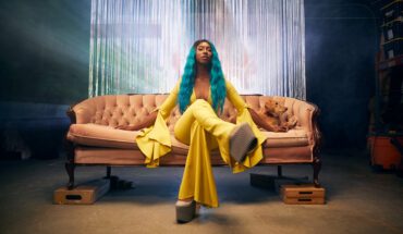 Domanique Grant press photo. Domanique is sitting on a vintage pink couch wearing a yellow jumpsuit