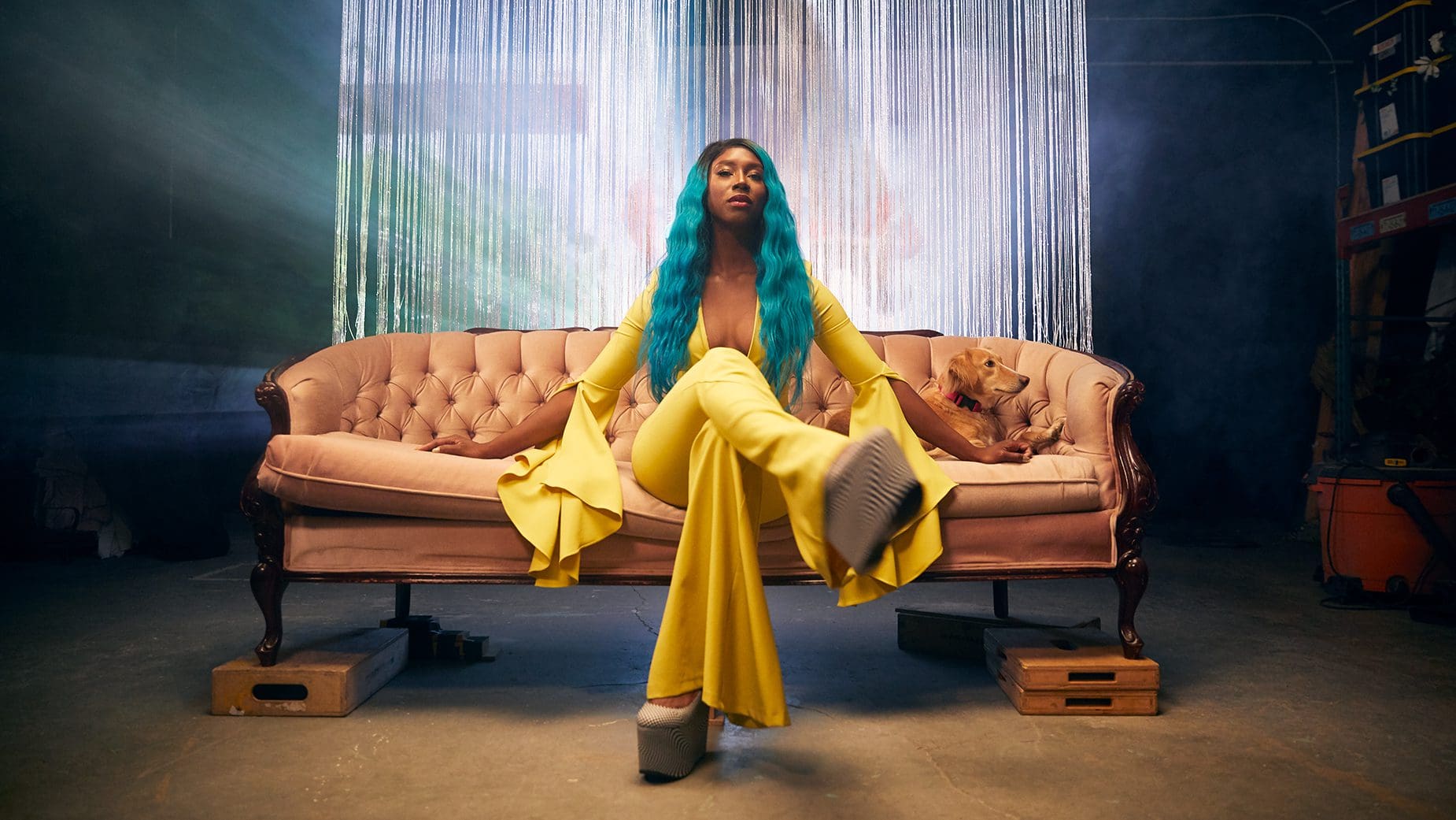 Domanique Grant Tackles Social Media Beauty Standards in Airbrush Domanique Grant press photo. Domanique is sitting on a vintage pink couch wearing a yellow jumpsuit