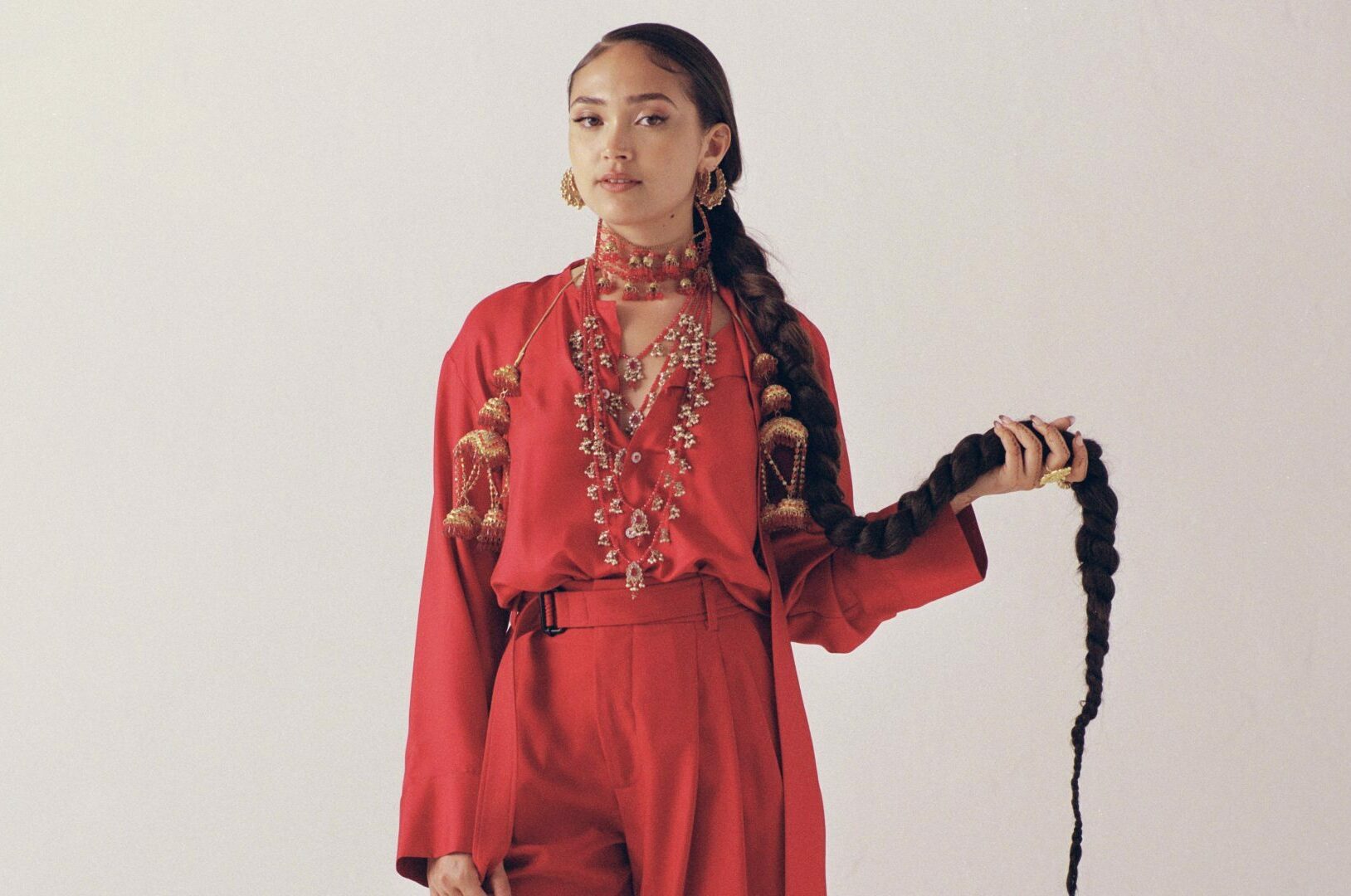 Joy Crookes press photo. Joy is waring an orange jumpsuit and standing in front of a beige backdrop.